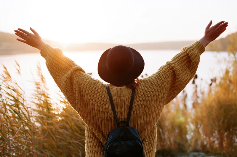 Woman with arms raised facing sunset over lake, wearing hat and sweater, backpack on back, surrounded by reeds