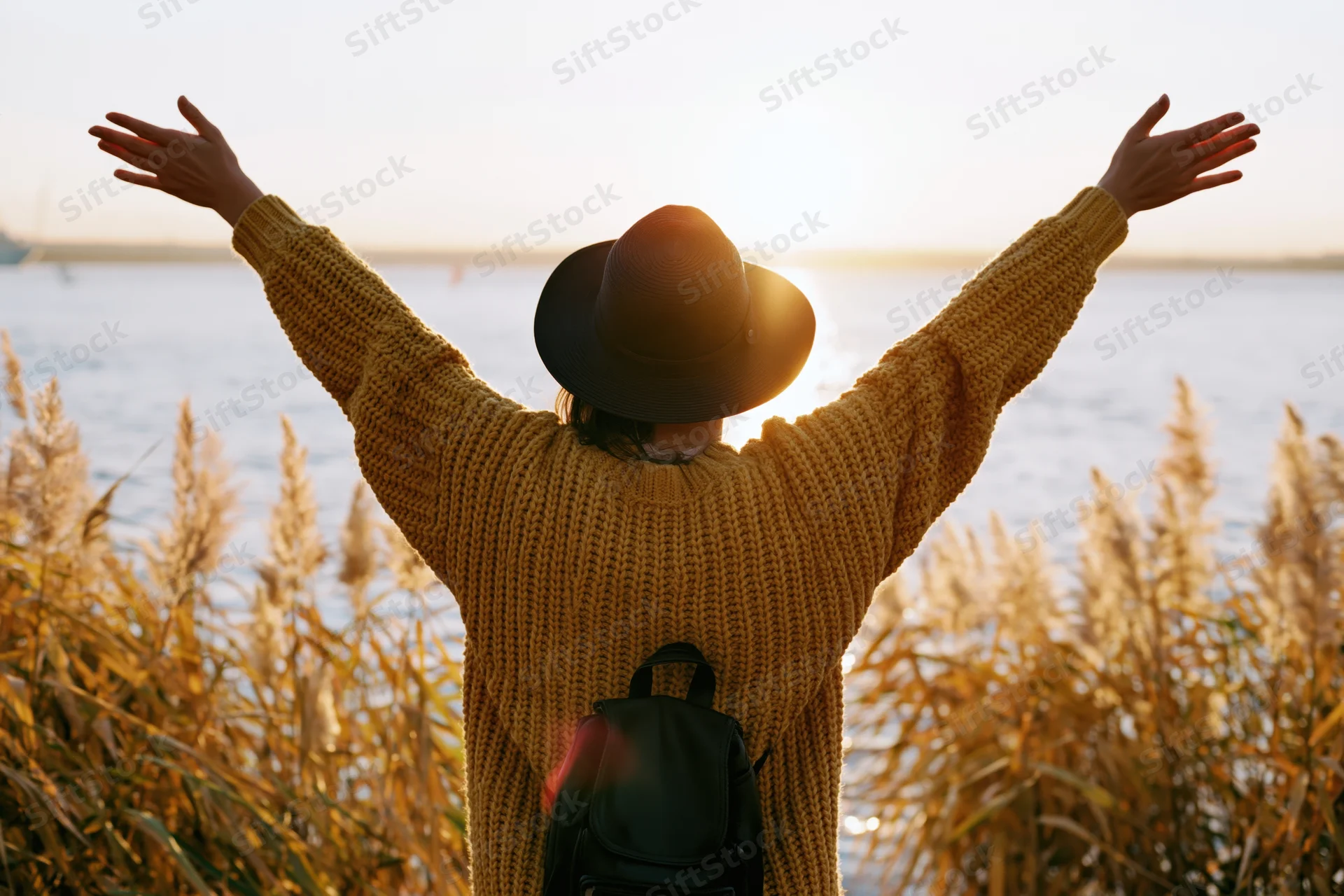 Woman with arms raised facing sunset over water surrounded by reeds