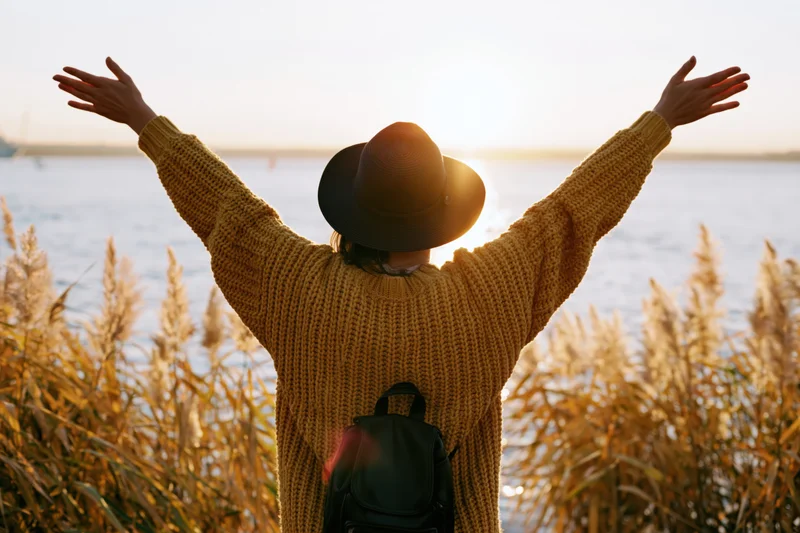 Woman with arms raised facing sunset over water surrounded by reeds