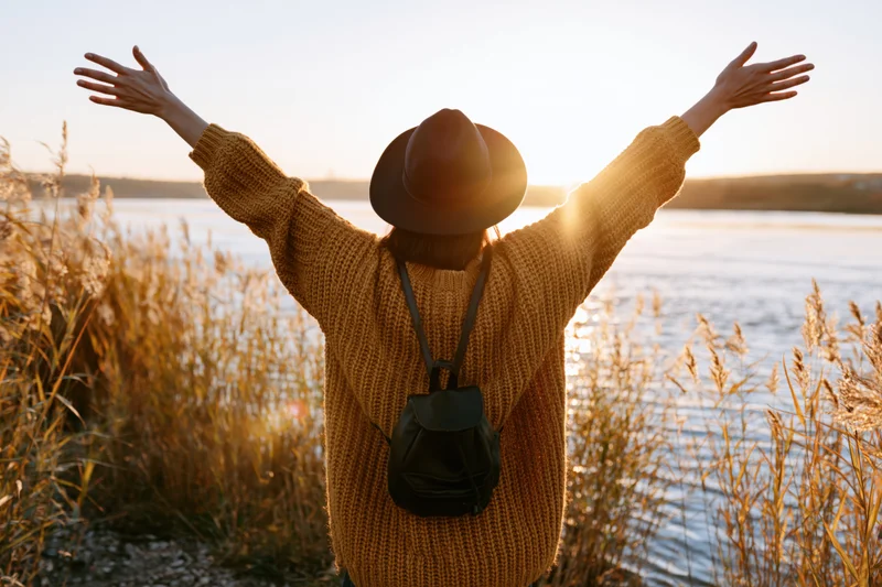 Woman with arms raised facing sunset by a lake surrounded by reeds