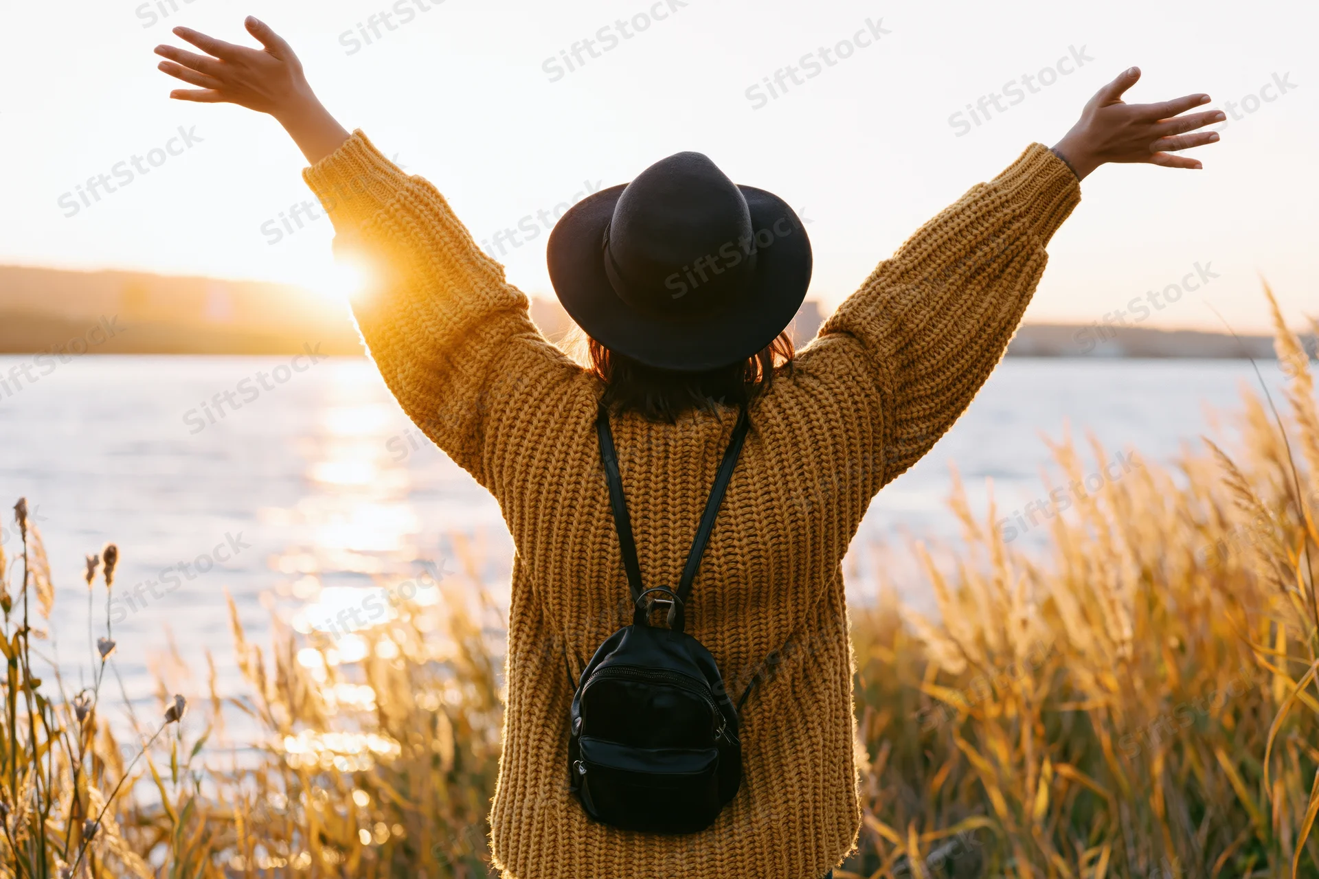 Woman with arms raised facing sunset over lake surrounded by tall grass