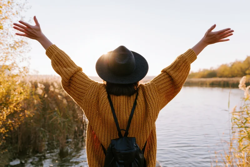 Woman with arms raised standing by a lake at sunset, back to camera, wearing a hat and sweater, surrounded by autumn foliage