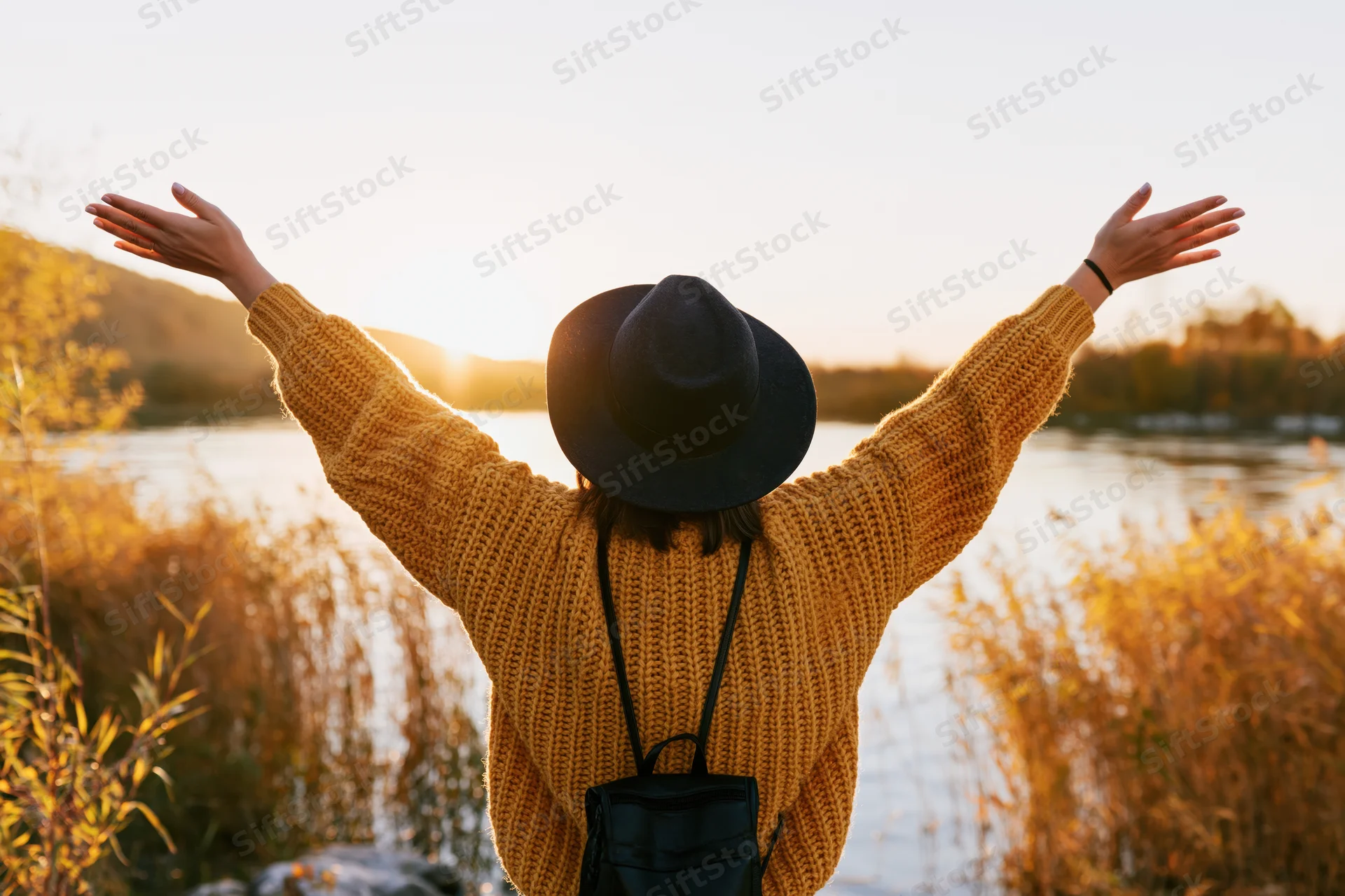 Woman with arms raised facing sunset by a lake surrounded by autumn reeds