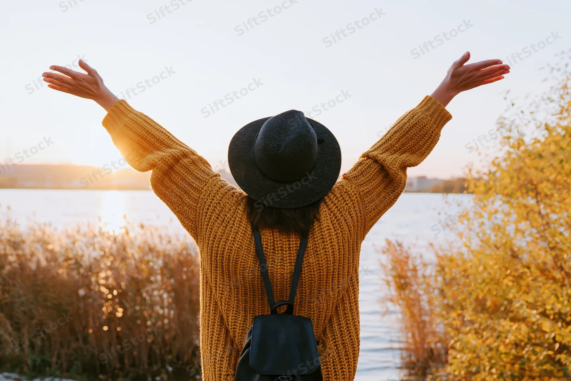 Woman with arms raised standing by a lake at sunset in autumn