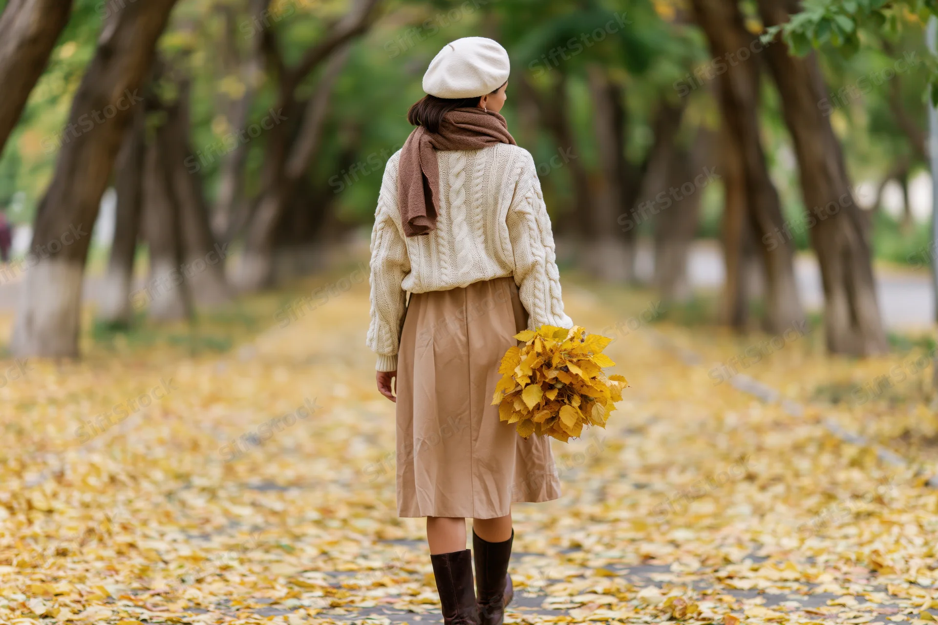 Woman walking down a leaf-covered park path in autumn holding a bundle of yellow leaves