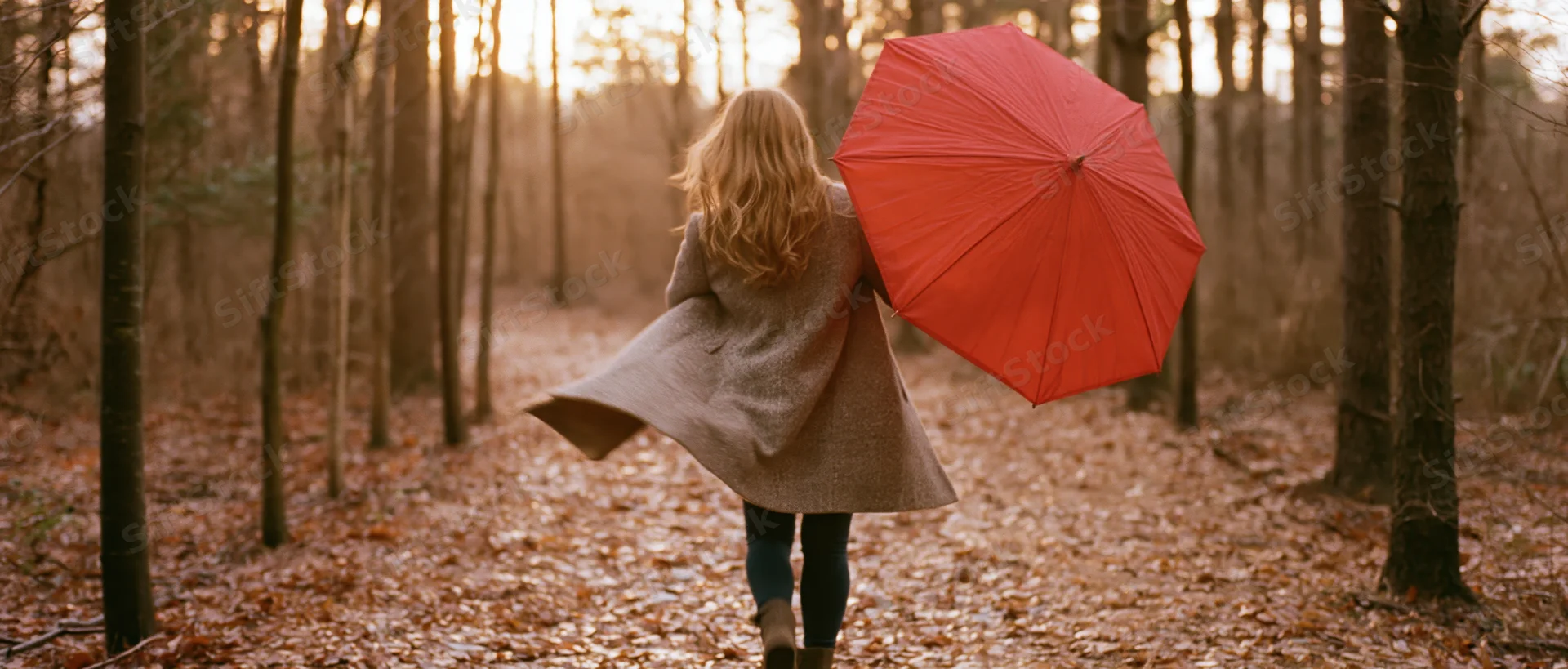 Woman walking away down a leaf-covered forest path holding a red umbrella at golden hour