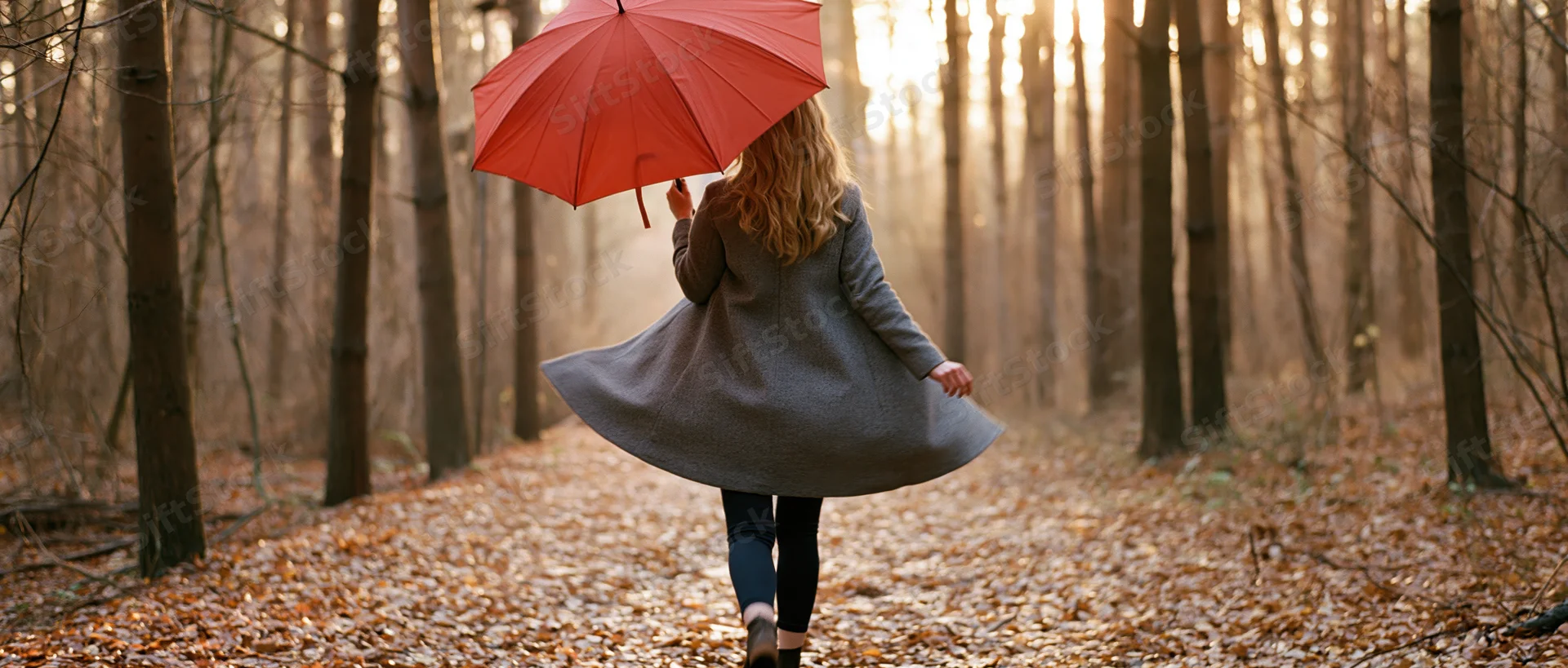 Woman walking away down a leaf-covered forest path holding a red umbrella