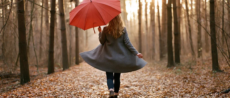 Woman walking away down a leaf-covered forest path holding a red umbrella
