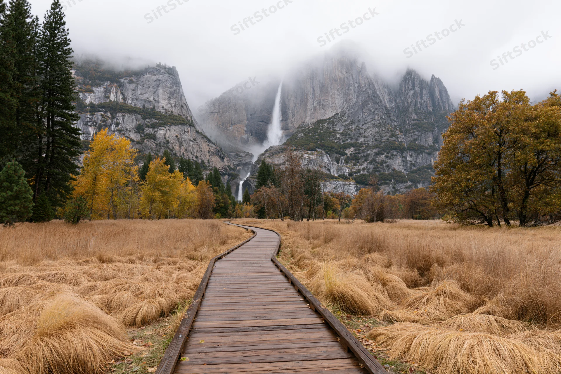 Wooden boardwalk path curving through golden autumn meadow with mountains and waterfall in background