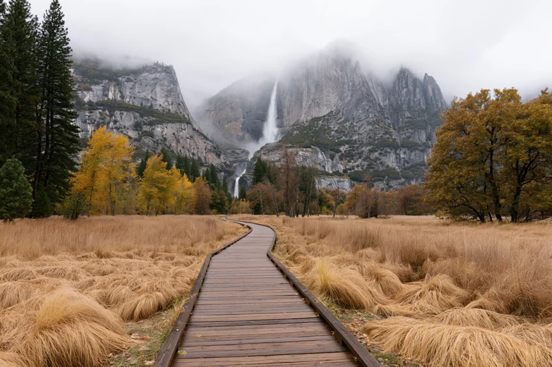 Wooden boardwalk path curving through golden autumn meadow with mountains and waterfall in background