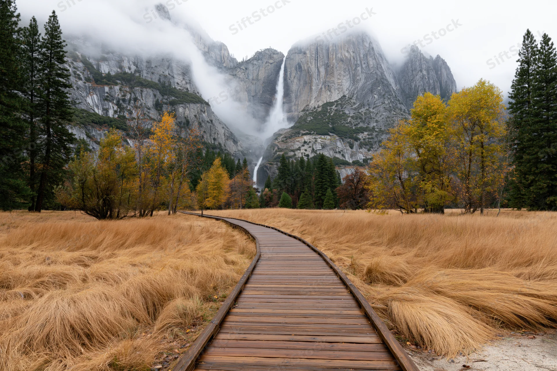 Wooden boardwalk path curving through golden autumn meadow toward misty mountains with waterfall