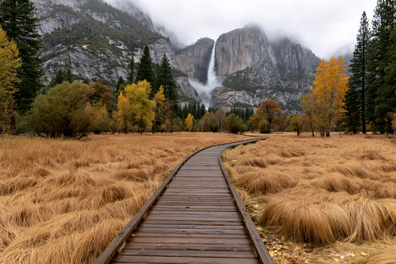 Wooden boardwalk path curving through golden autumn meadow with mountains and waterfall in background