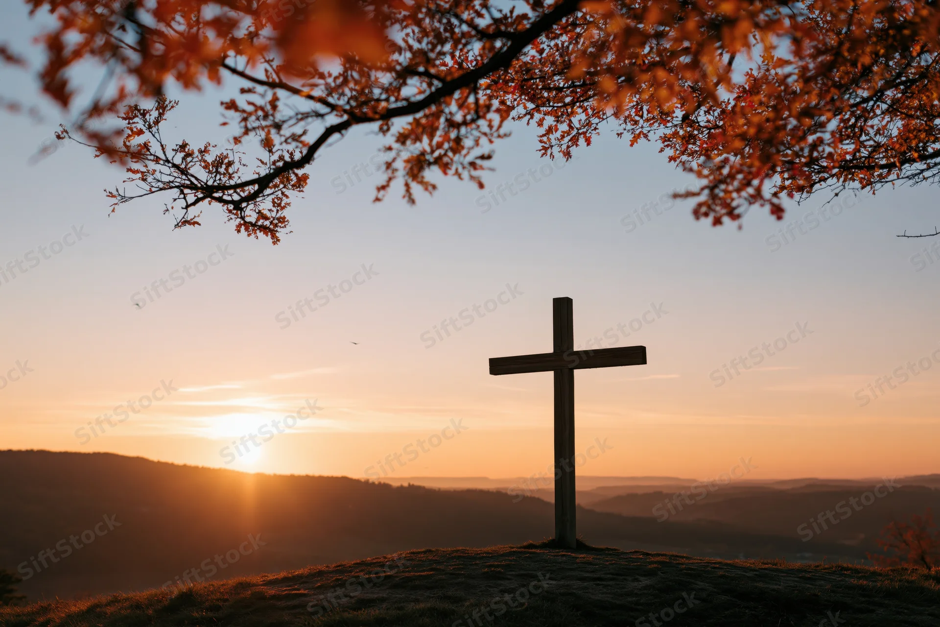 Wooden cross silhouetted on a hilltop at sunset with autumn leaves framing the scene