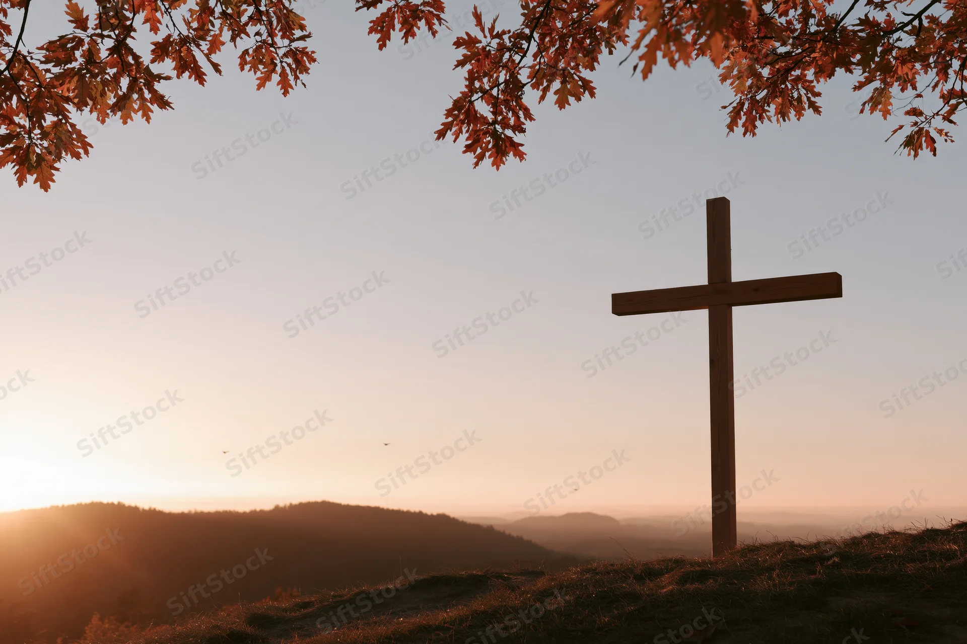 Wooden cross silhouetted on a hilltop at sunset with autumn leaves framing the sky