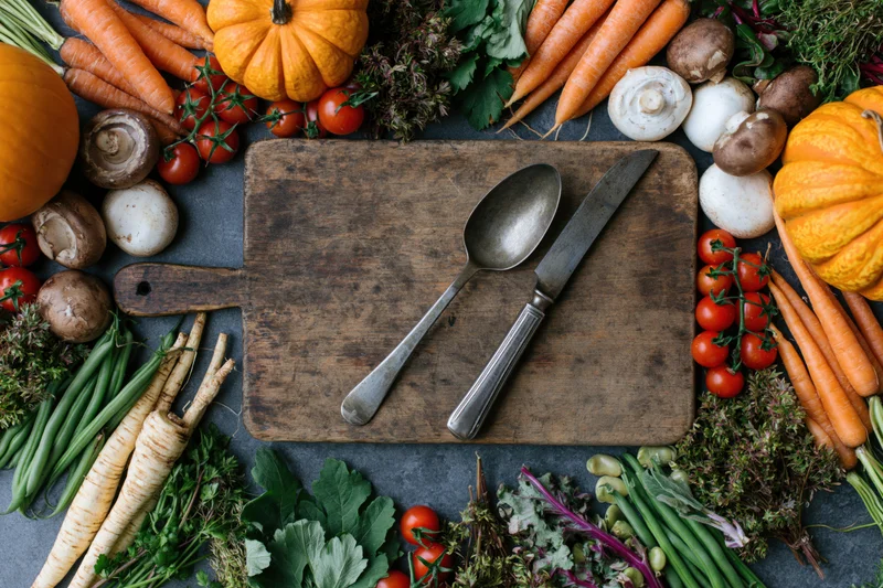 Fresh autumn vegetables arranged around a wooden cutting board with knife and spoon on dark slate surface