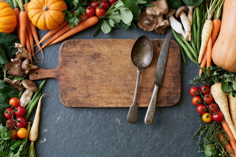 Wooden cutting board with knife and spoon surrounded by fresh vegetables on dark slate surface