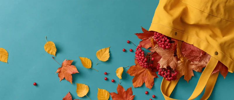 Yellow canvas tote bag overflowing with autumn leaves and red rowan berries on teal background