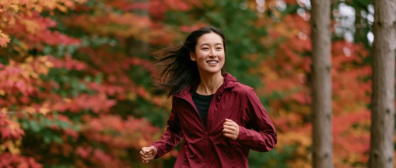 Young asian woman jogging through autumn forest trail smiling with windblown hair