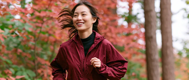 Young asian woman jogging through autumn forest trail smiling with wind in her hair