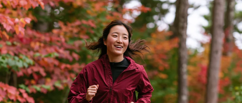 Young asian woman jogging through autumn forest trail smiling with wind in her hair