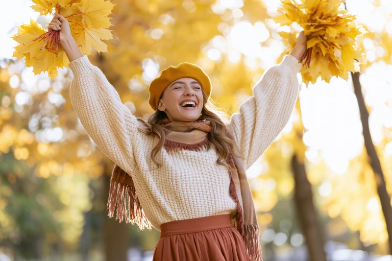 Woman laughing with arms raised holding bunches of yellow maple leaves in autumn park