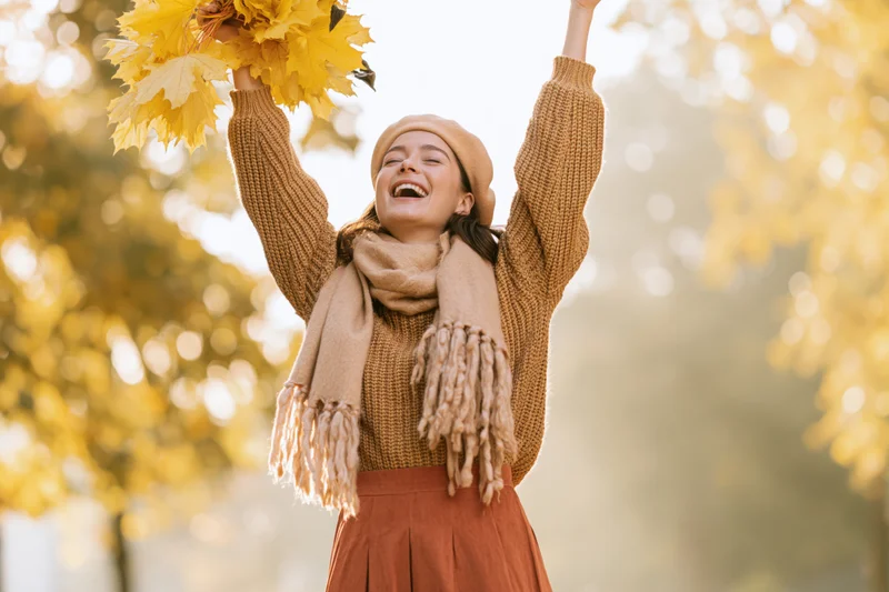Woman laughing with arms raised holding yellow leaves in autumn park