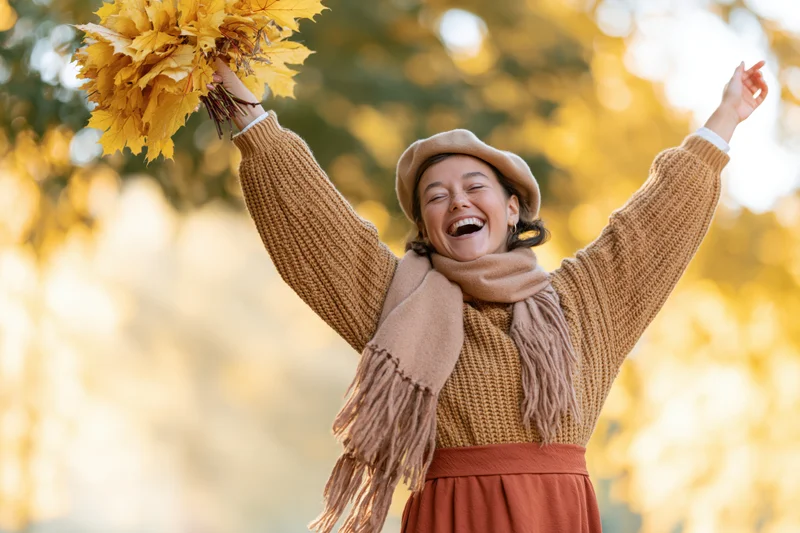 Woman laughing with arms raised holding bundle of yellow autumn leaves outdoors