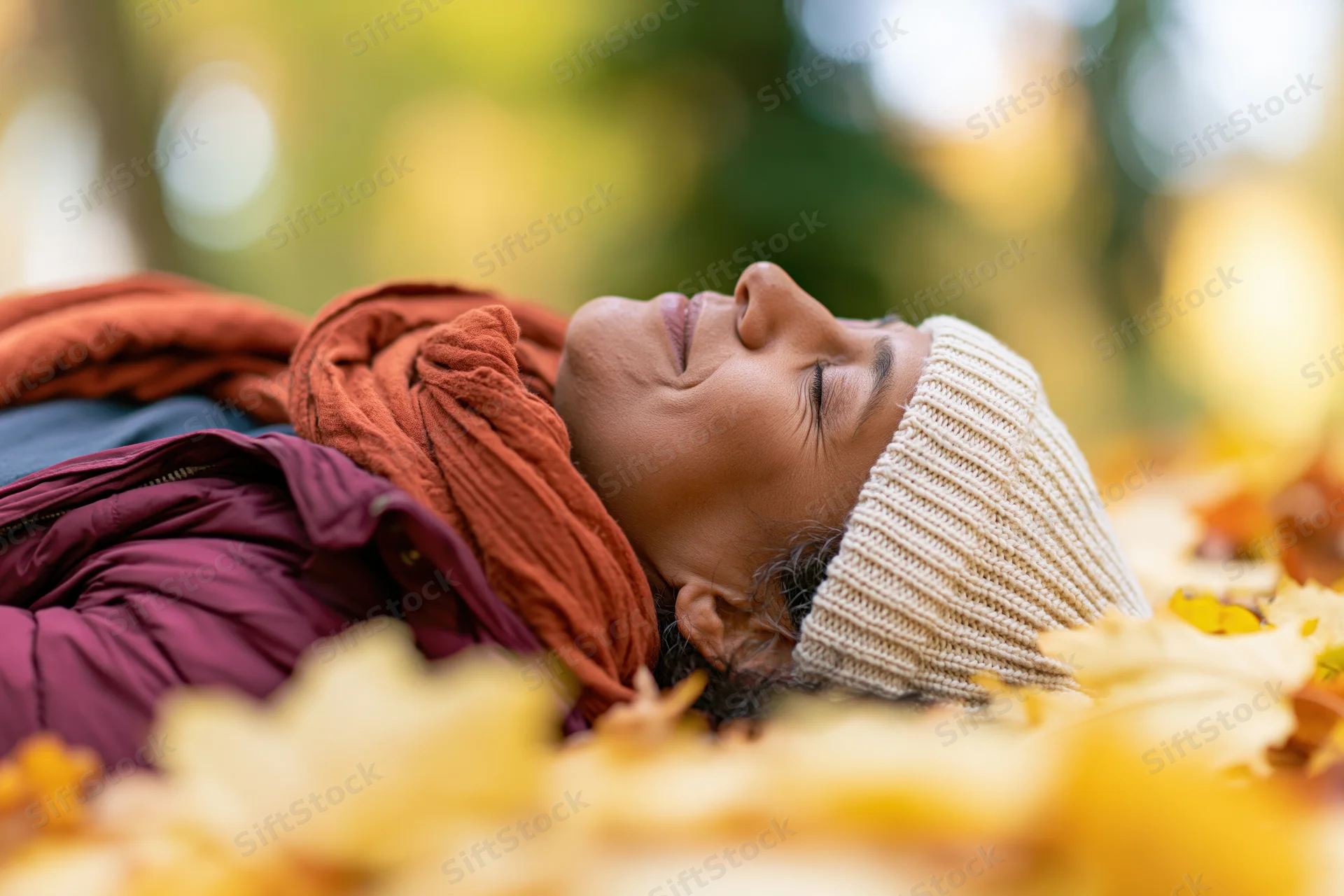 Woman lying on back in autumn leaves with eyes closed and smiling, outdoors in park