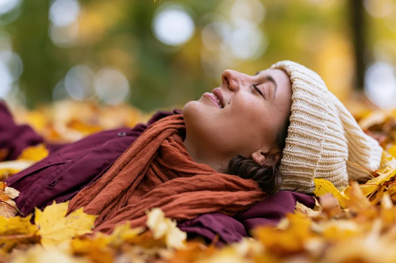 Woman lying on back in pile of autumn leaves in park, eyes closed, wearing beanie and scarf