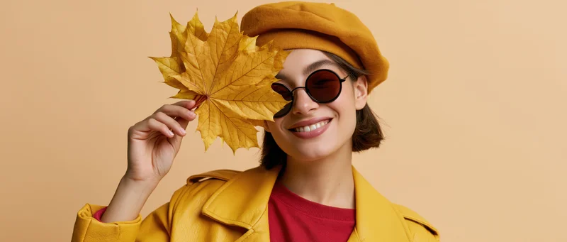 Woman smiling while holding a golden maple leaf in front of her face, wearing yellow coat and beret against beige background