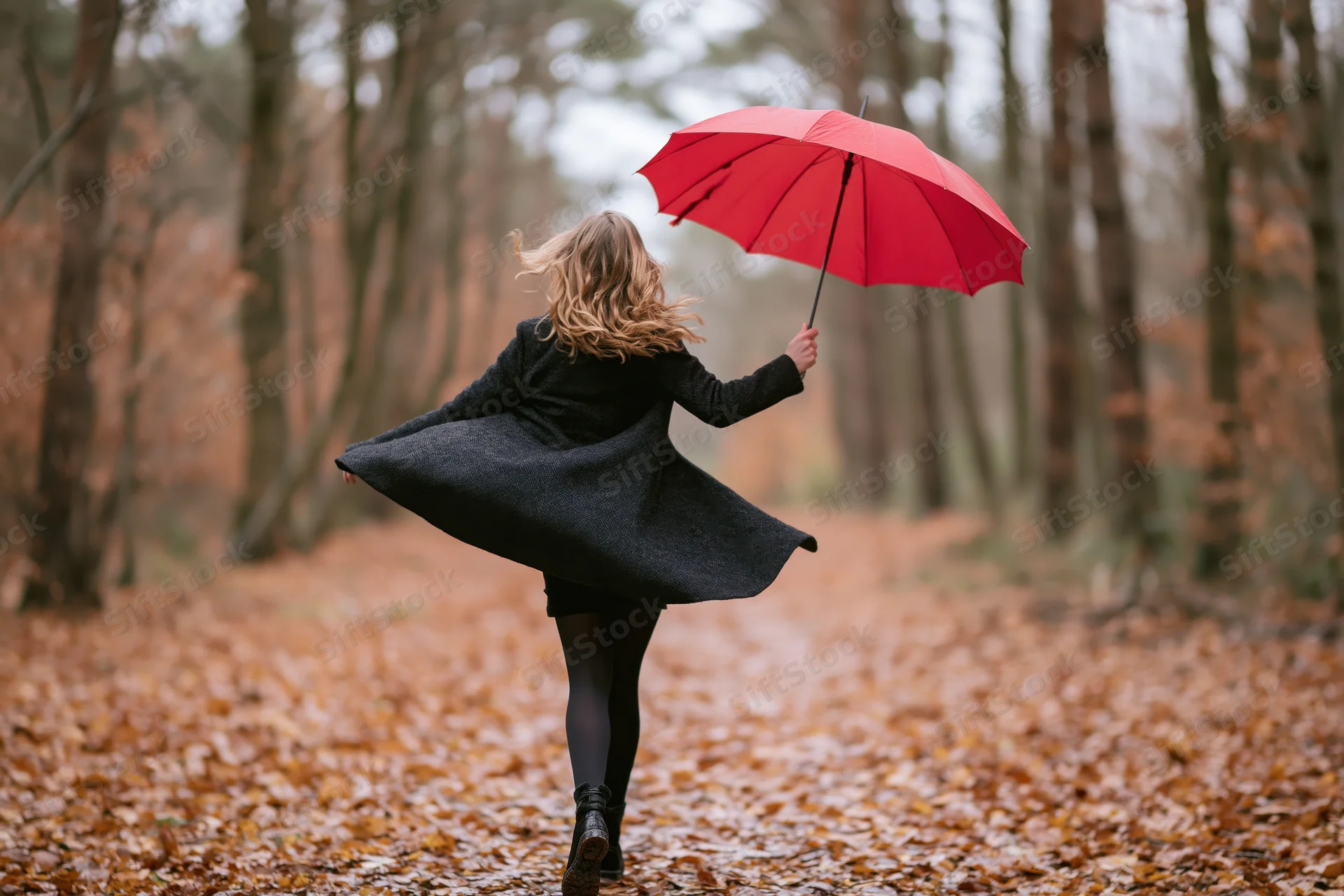 Woman twirling with red umbrella on leaf-covered forest path in autumn