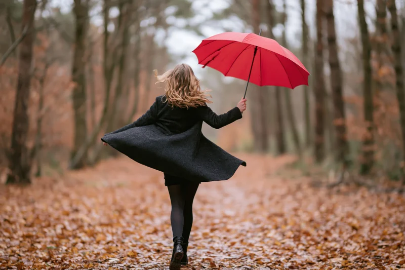 Woman twirling with red umbrella on leaf-covered forest path in autumn