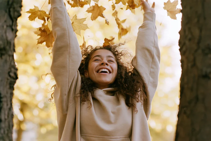 Young woman laughing with arms raised among falling autumn maple leaves in a sunlit park