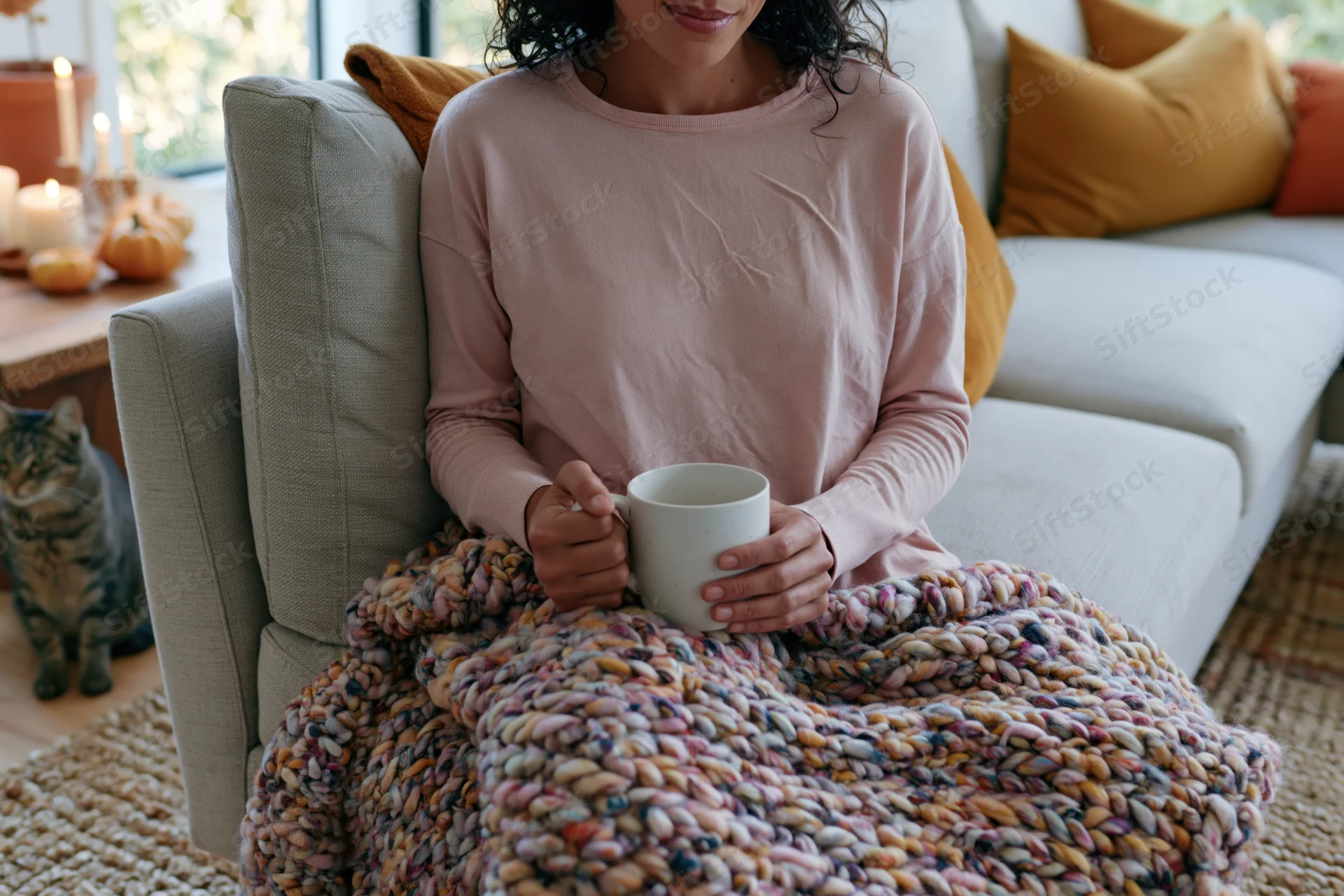 Woman sitting on sofa wrapped in knitted blanket holding mug in living room with cat and autumn decor
