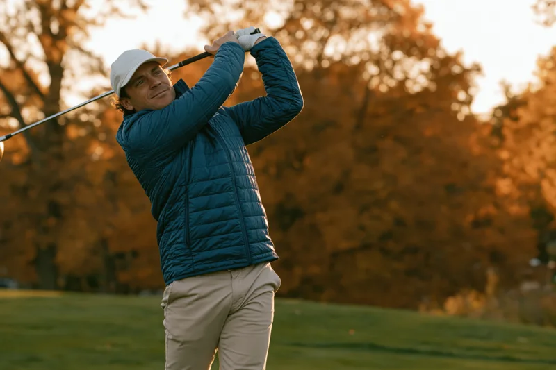 Man swinging golf club on a grassy course with autumn trees in the background