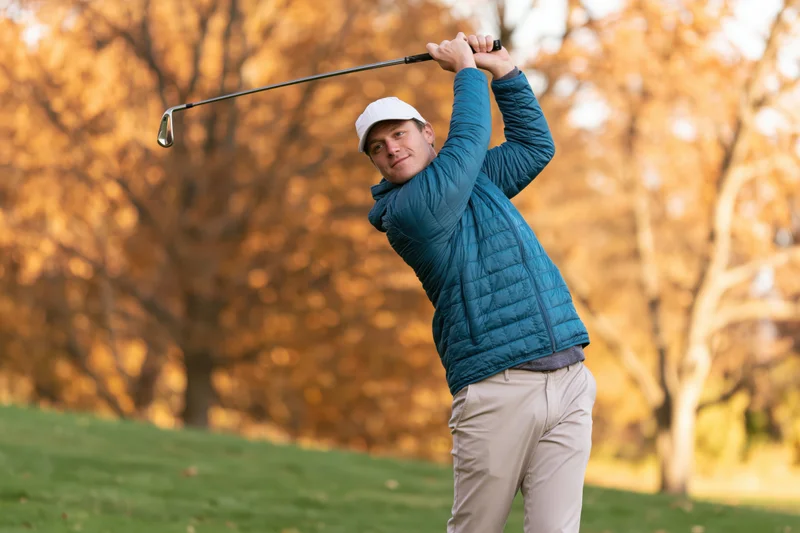 Man swinging golf club on grassy course with autumn trees in background