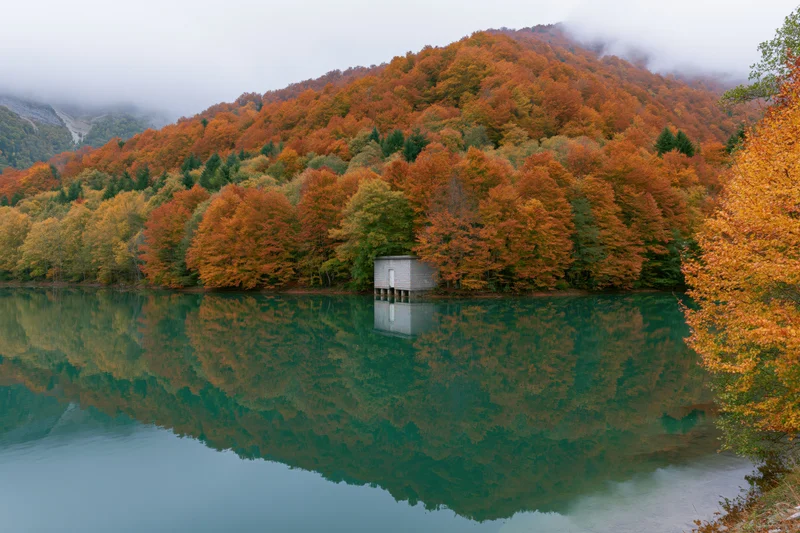 Calm mountain lake reflecting autumn forest and small stone cabin on shore