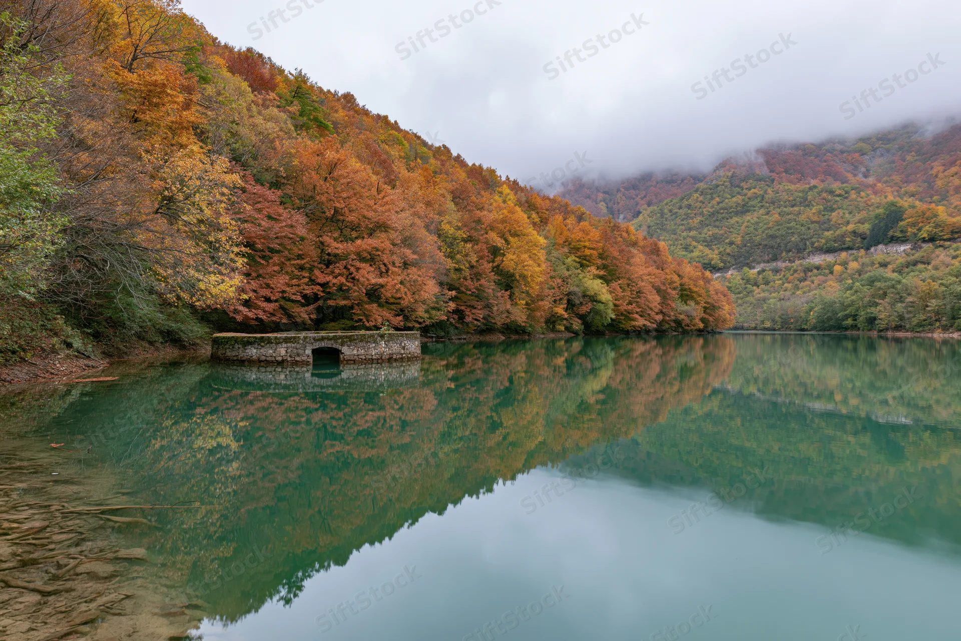 Calm mountain lake reflecting autumn forest and stone structure along the shore