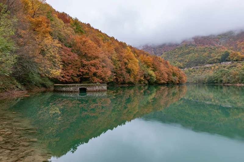 Calm mountain lake reflecting autumn forest and stone structure along the shore