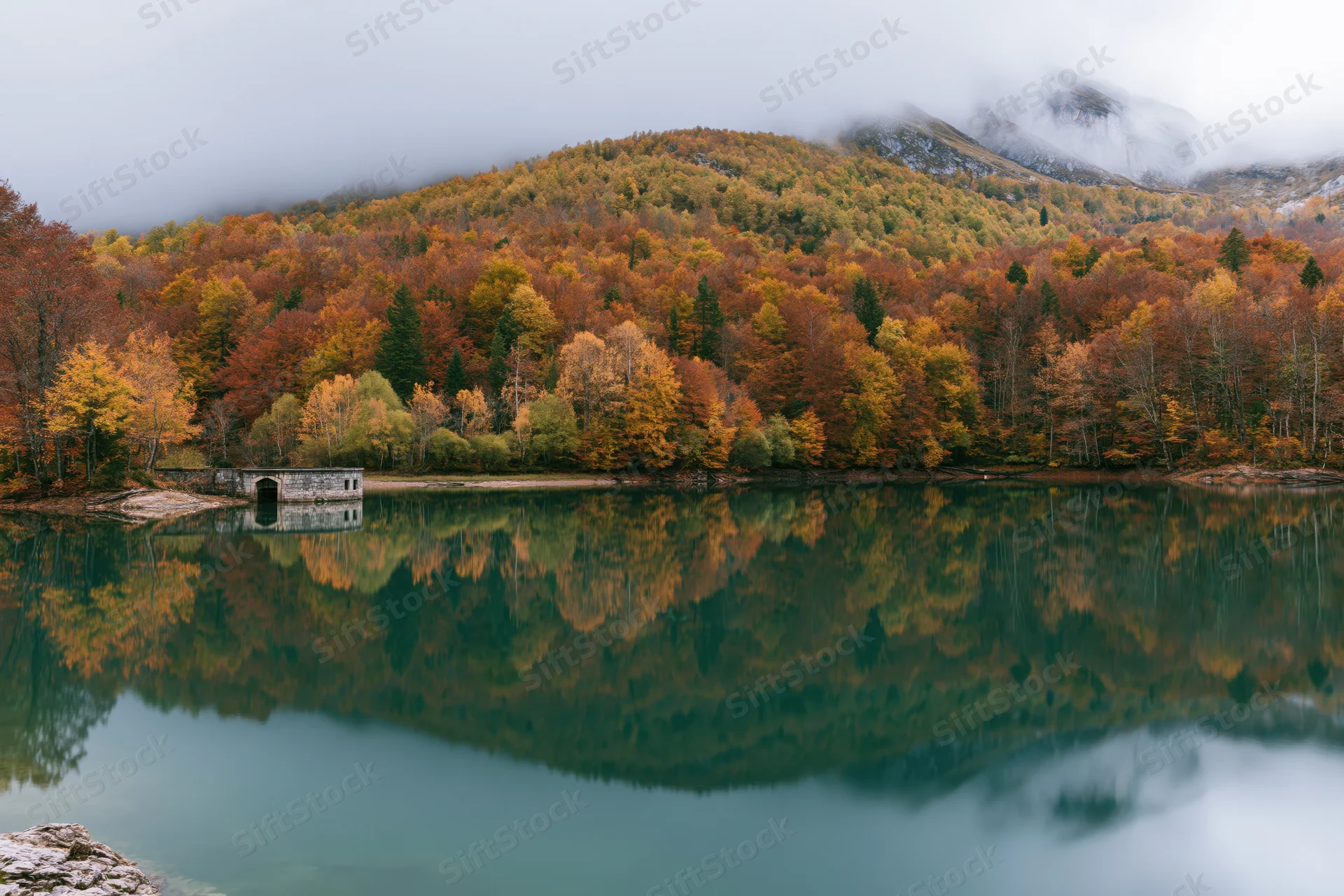 Calm mountain lake reflecting autumn forest and stone structure under misty sky