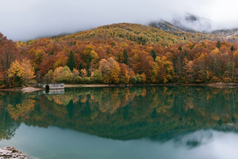 Calm mountain lake reflecting autumn forest and stone structure under misty sky