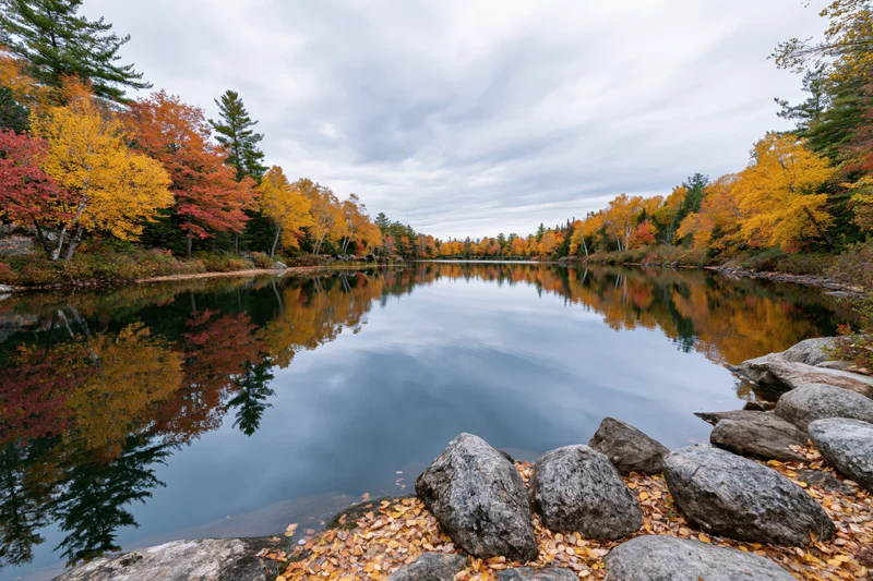 Calm lake reflecting autumn trees along rocky shore under cloudy sky