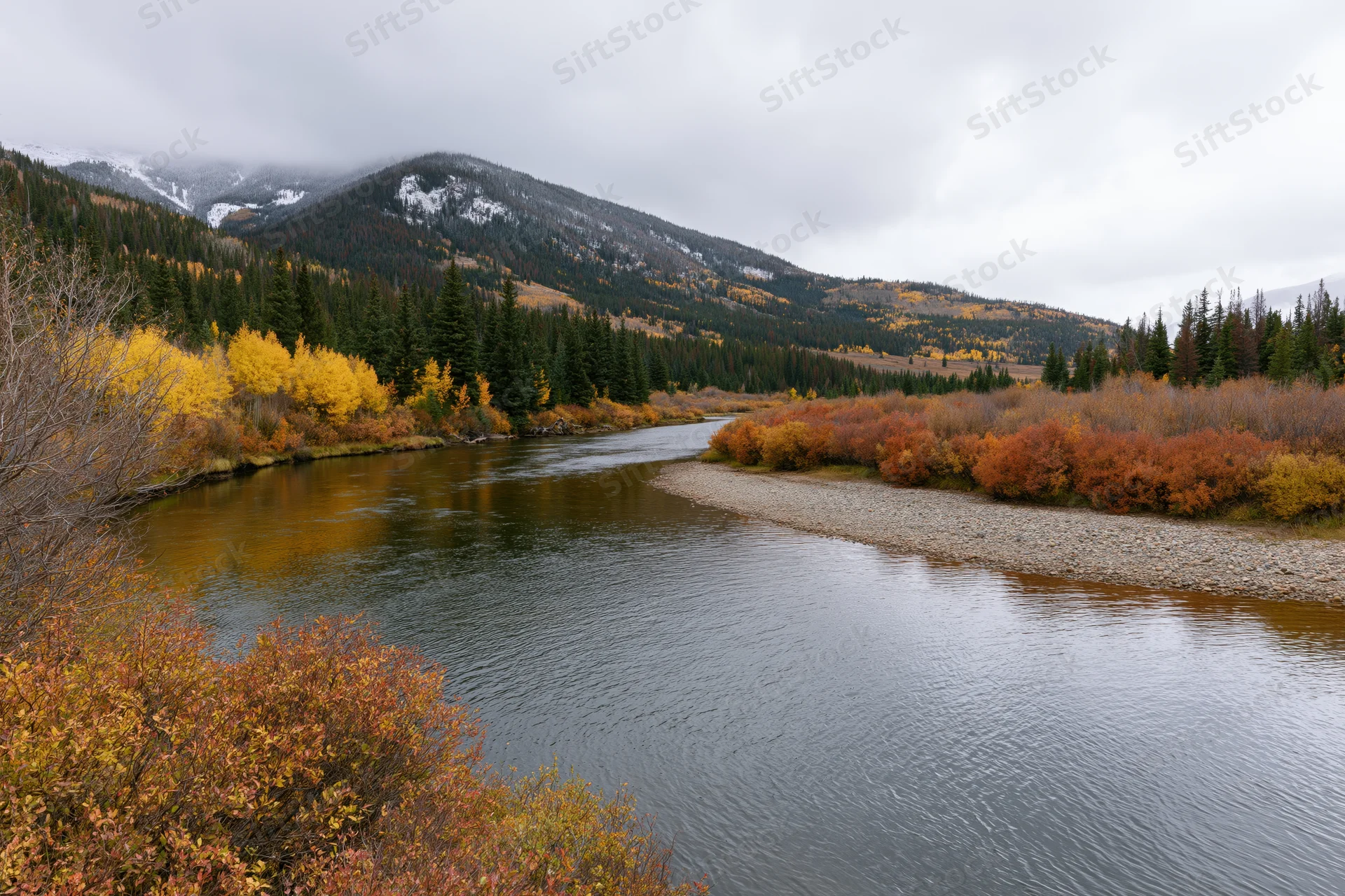 Calm river winding through a mountain valley in autumn with colorful trees and snow-capped peaks under an overcast sky