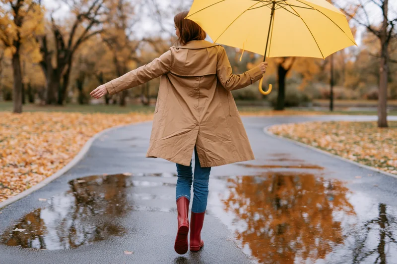Woman walking on wet park path with yellow umbrella, autumn leaves scattered around