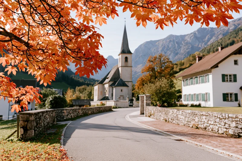 Winding road through a small alpine village with autumn foliage framing a church and mountains in the background
