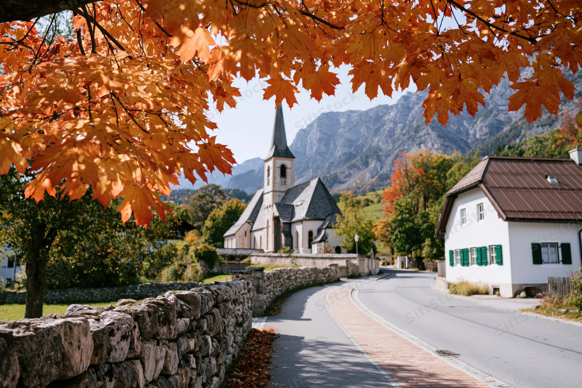 Winding road through alpine village framed by autumn maple leaves with church and mountains in background