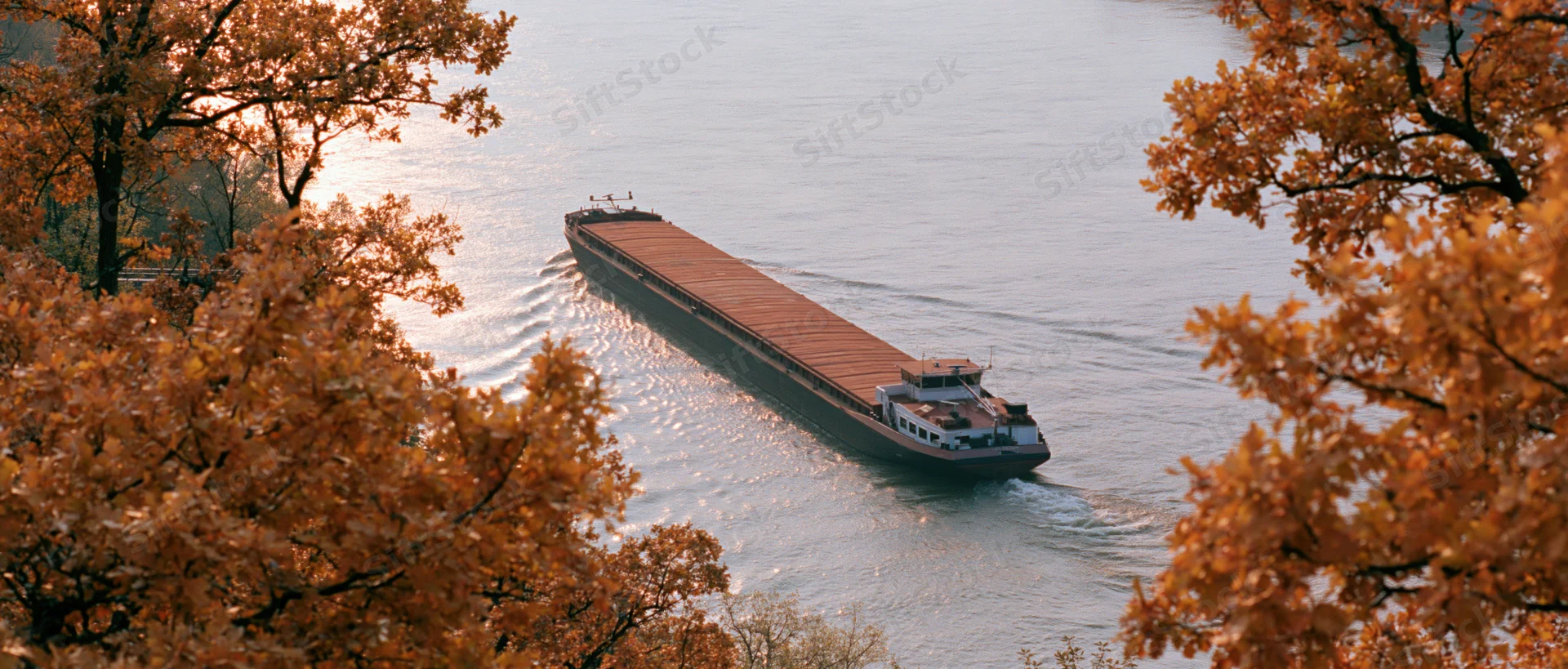 Flat-bottomed cargo barge moving slowly along a wide river framed by autumn trees