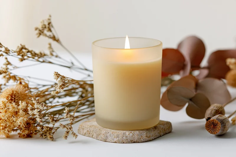 Lit cream soy candle in frosted glass jar on stone coaster surrounded by dried flowers and botanicals