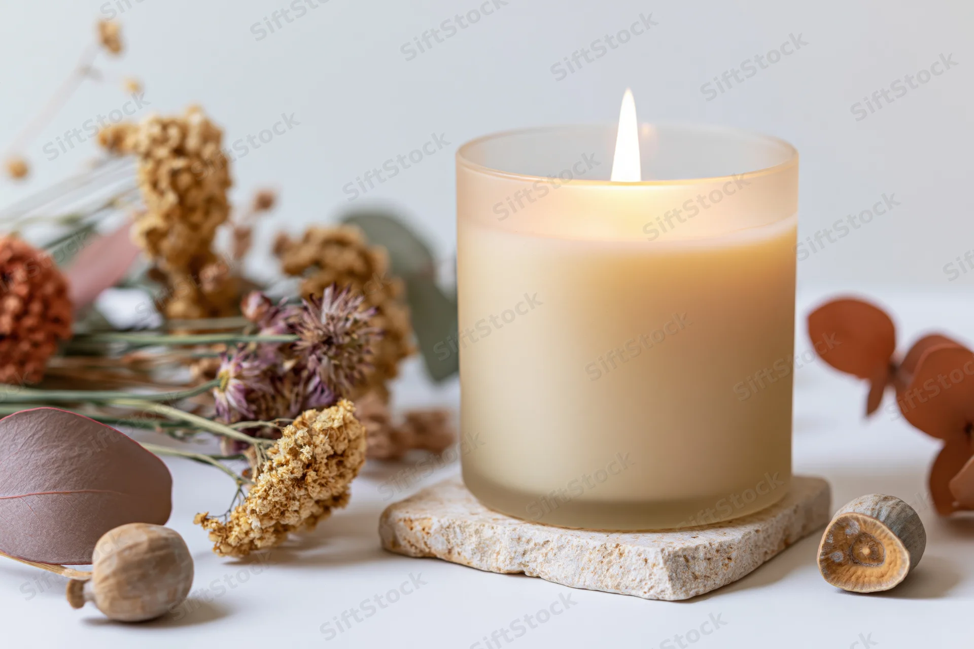Lit cream soy candle in frosted glass jar on stone coaster surrounded by dried flowers and acorns on white surface