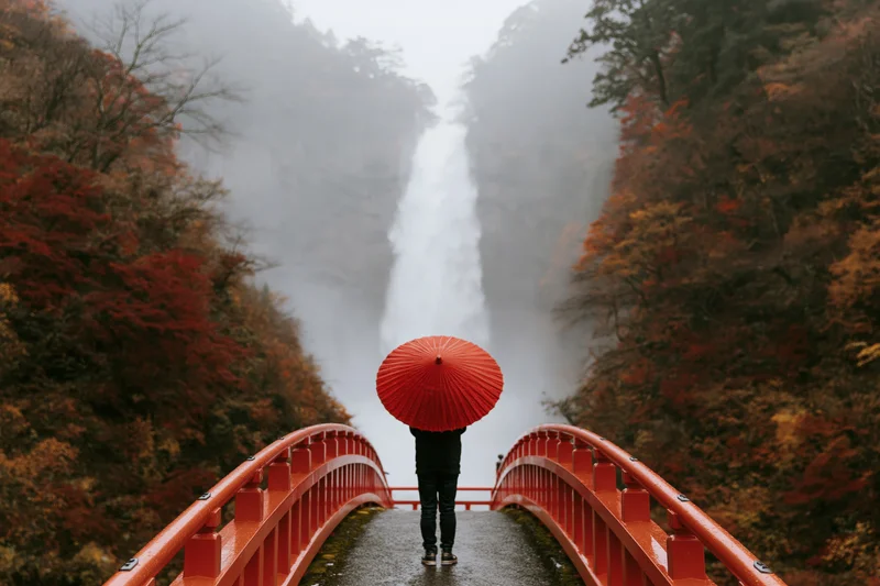 Person holding red paper umbrella on arched bridge facing waterfall in autumn forest